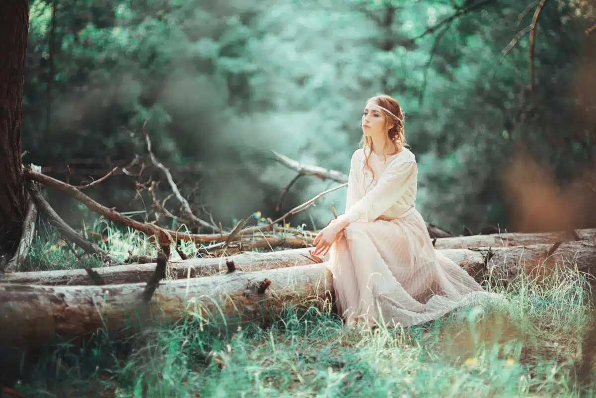 Beautiful artistic photo ginger girl in white dress sitting on a tree in the woods.