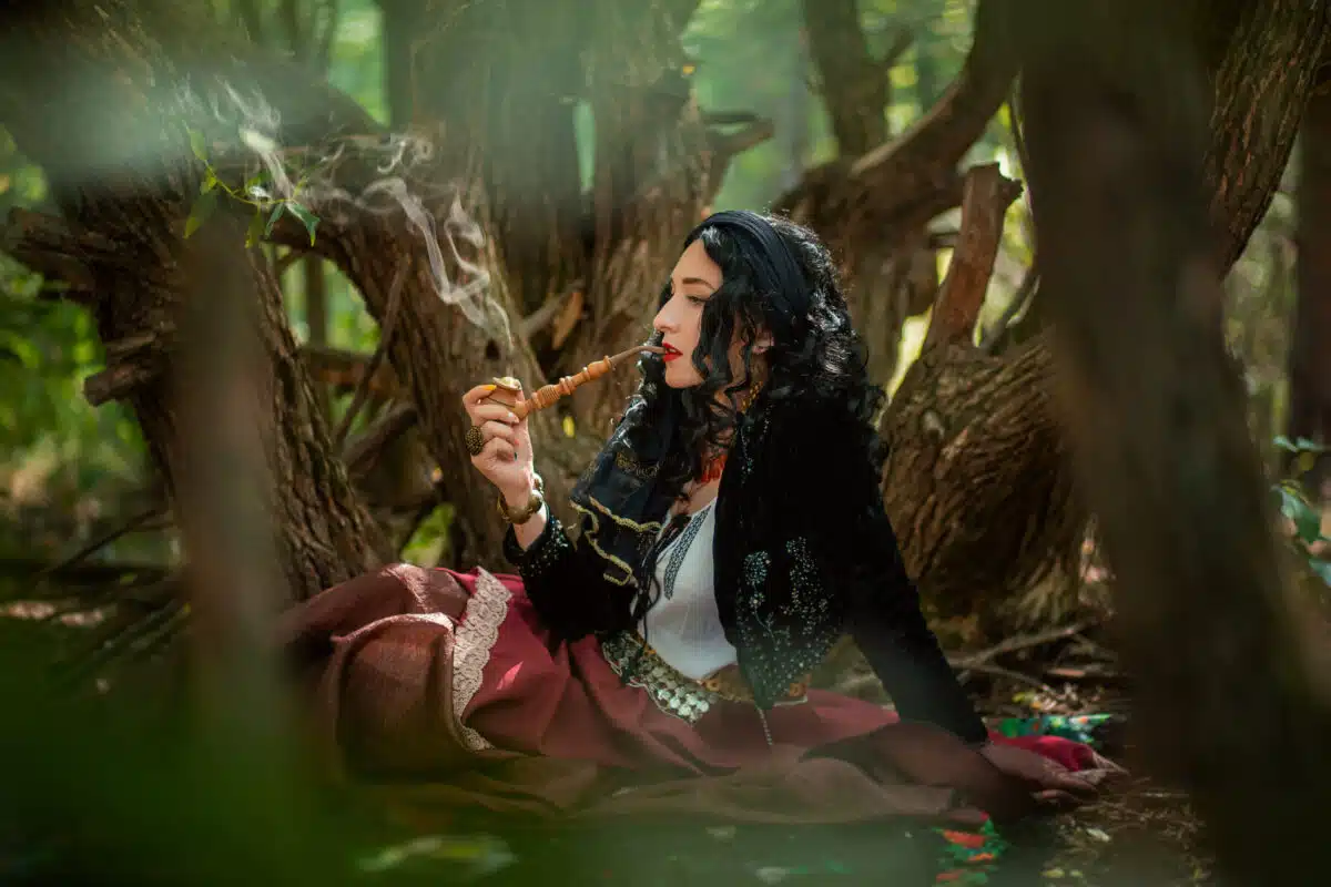 A gypsy woman in the forest with a wooden smoking pipe in her hands. The black-haired witch conjures in the forest. Women's Magic.