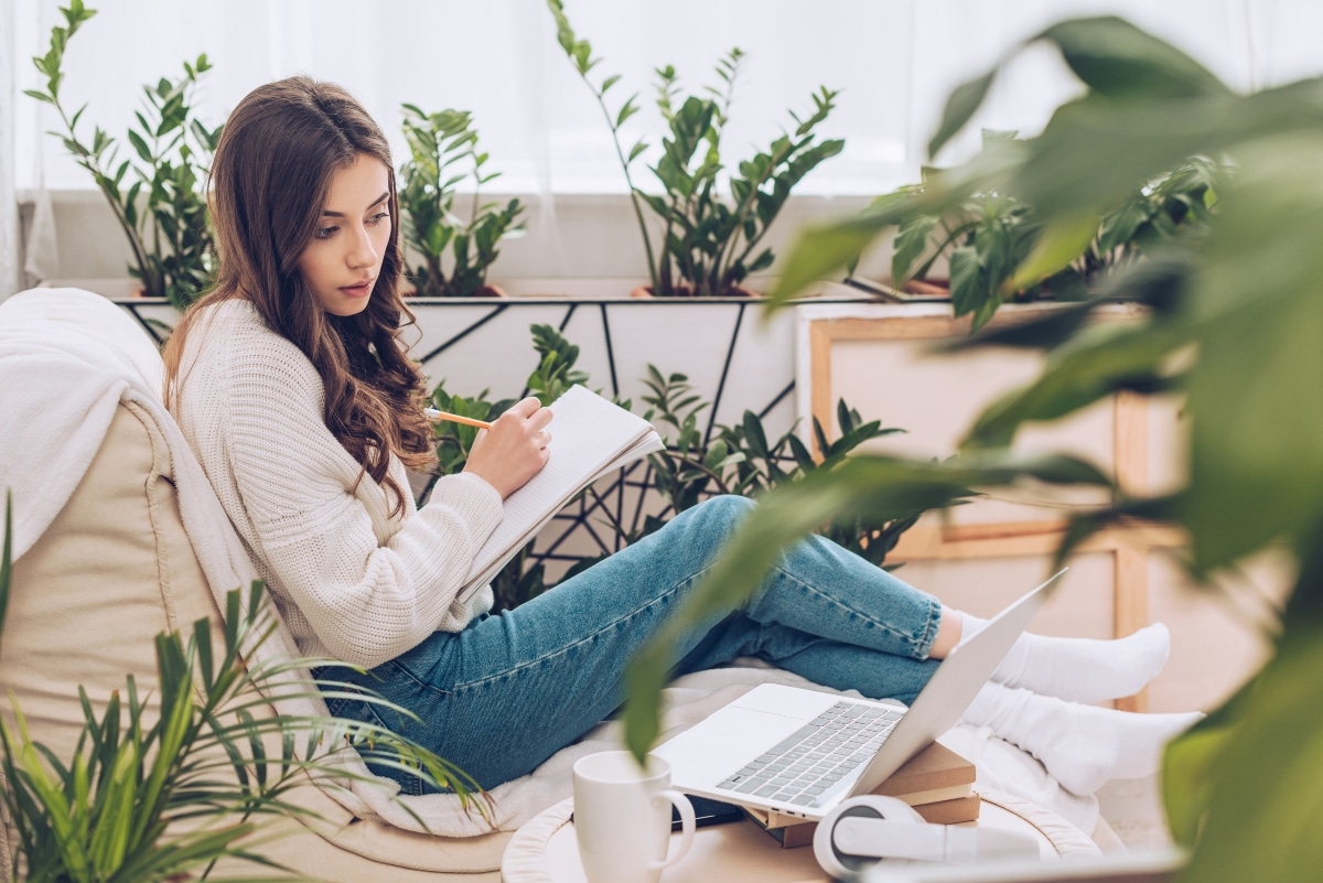 selective focus of attentive young woman writing using laptop and writing in notebook while sitting near green plants at home