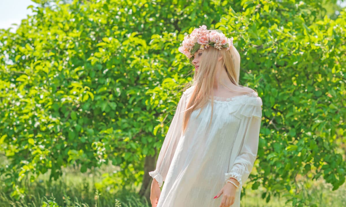 a lady in a long-sleeved white dress and flower head wreath standing in the garden