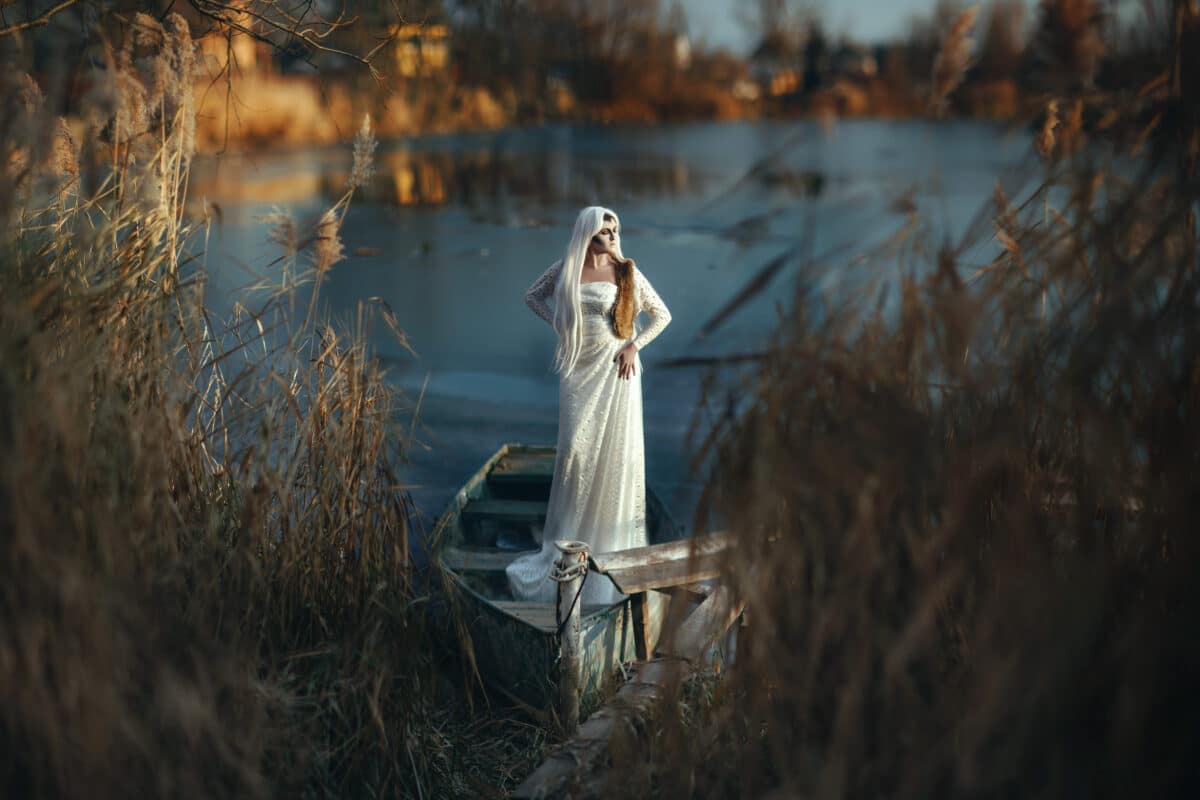 nymph dressed in white standing on a boat on an icy lake behind tall grass