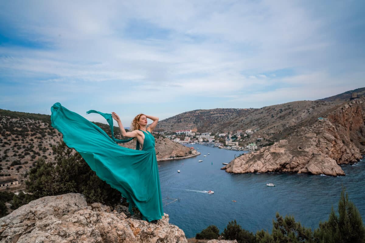 A woman in an emerald dress, with long silk wings in a flowing dress standing on the edge of a mountain with ocean view