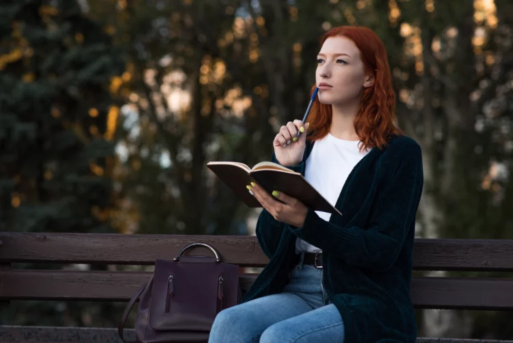 A young and attractive redhead sitting on a bench, holding a notepad and thinking.