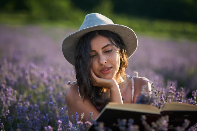 Beautiful girl in hat sitting in purple lavender field and read book.