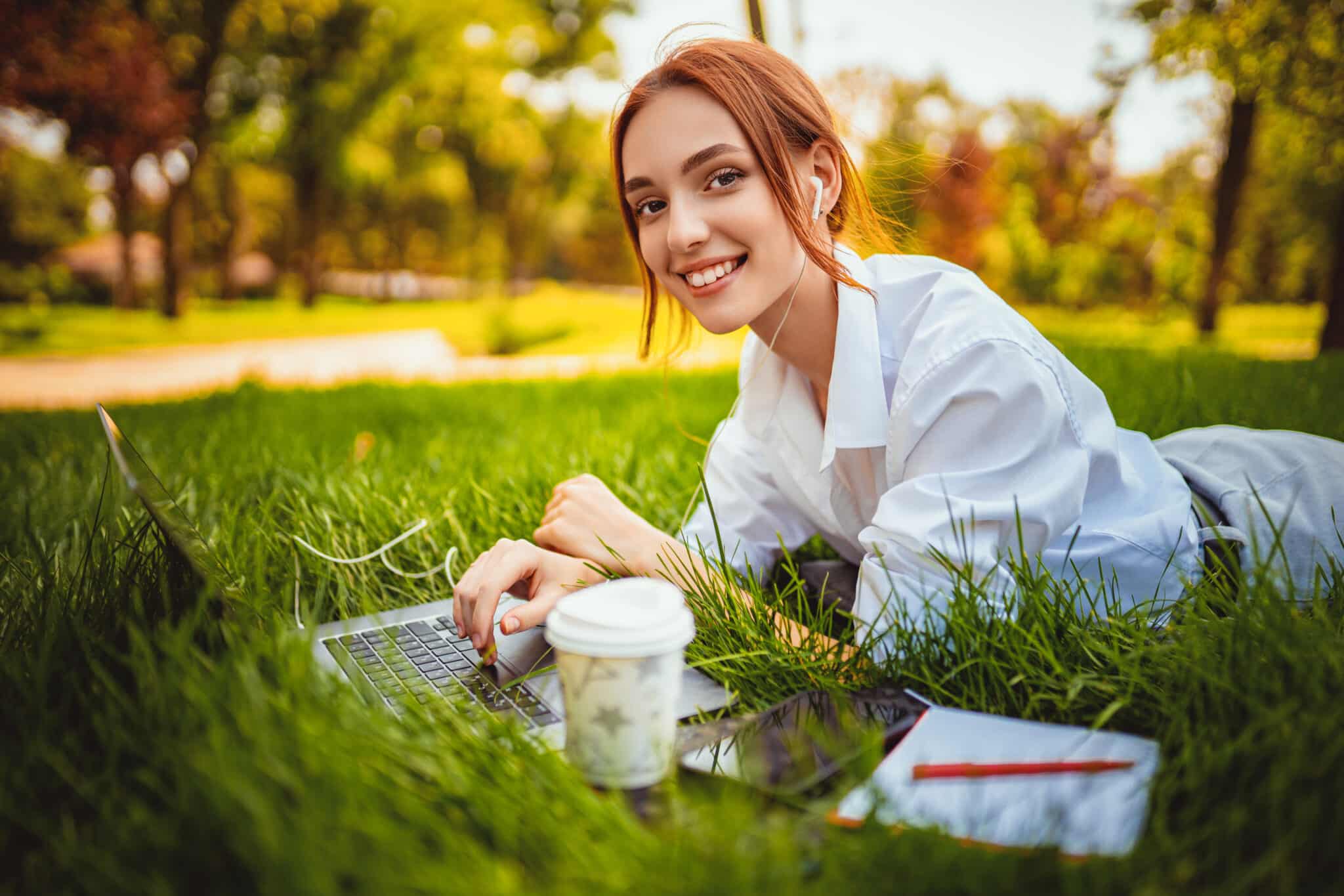 beautiful smiling female student uses laptop pc while lying on the grass outdoors. Education concept