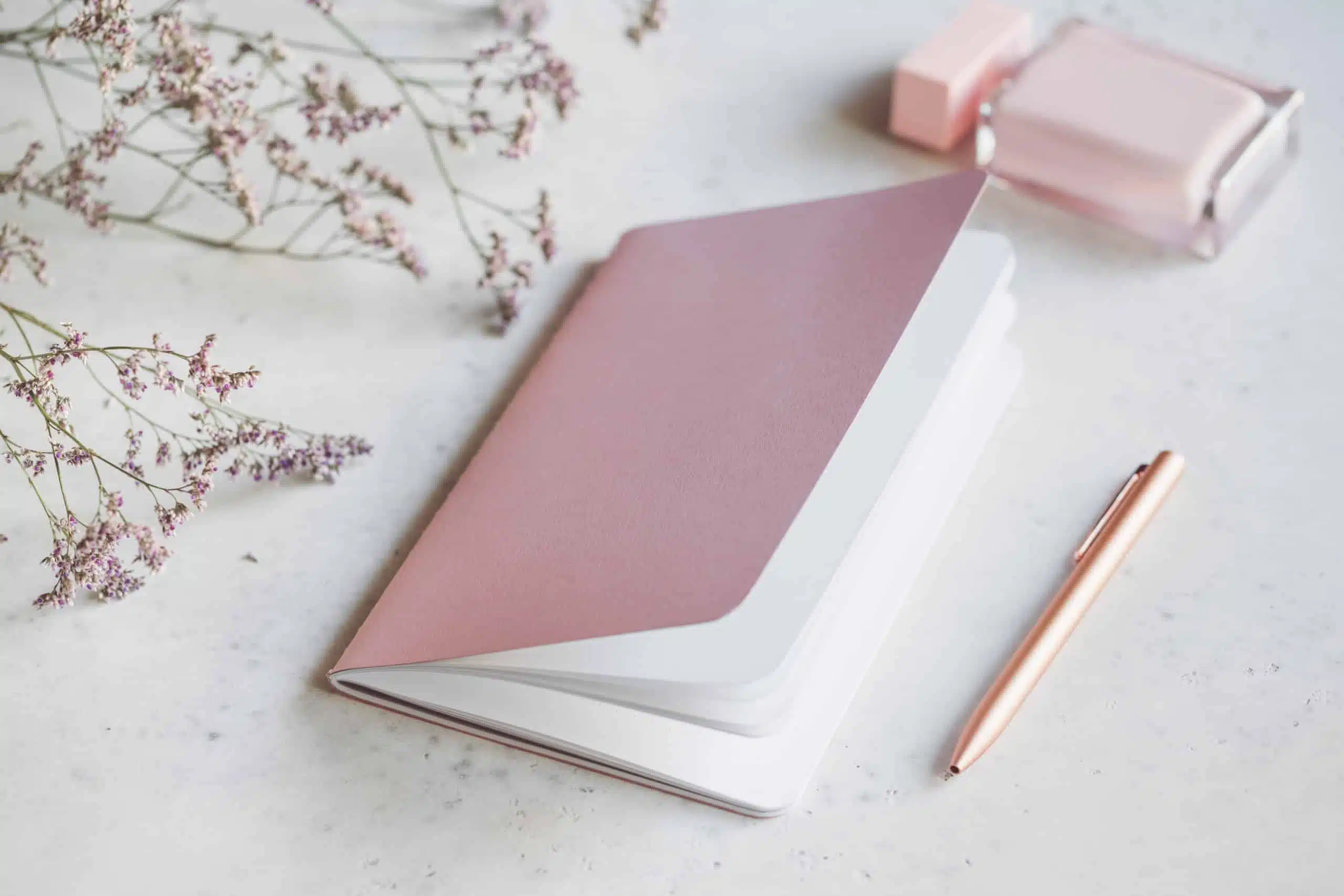 Empty pink notebook with golden pen on white table surrounded by flowers and fragrance.
