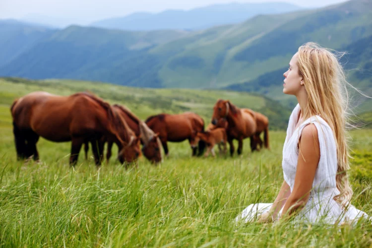 Woman sitting on mountain top, with brown horses grazing in the background
