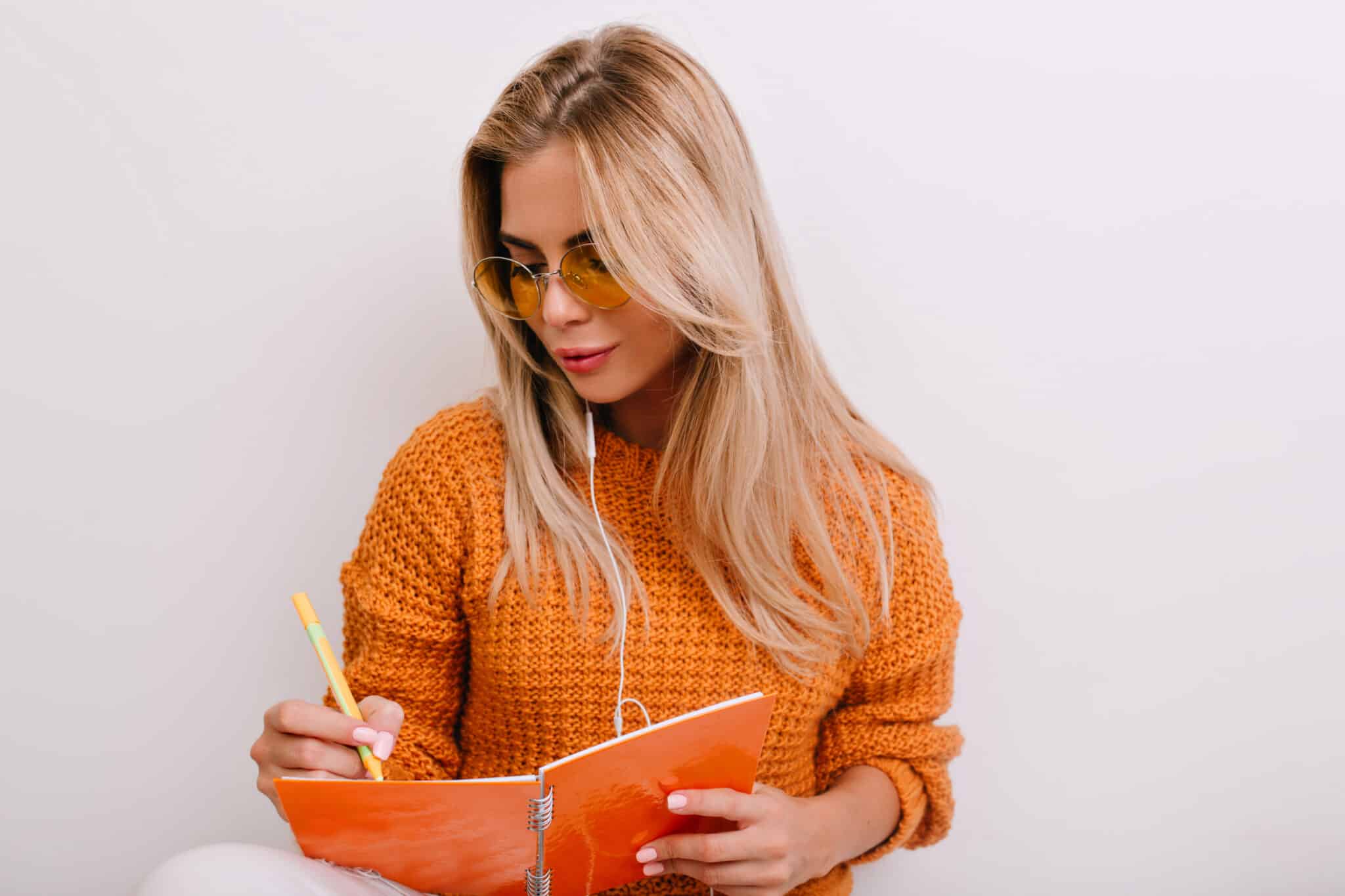 Lovely blonde woman with elegant manicure writing something in colorful notebook with gently smile. Indoor portrait of stunning lady in wool sweater drawing in sketchbook and listening music..
