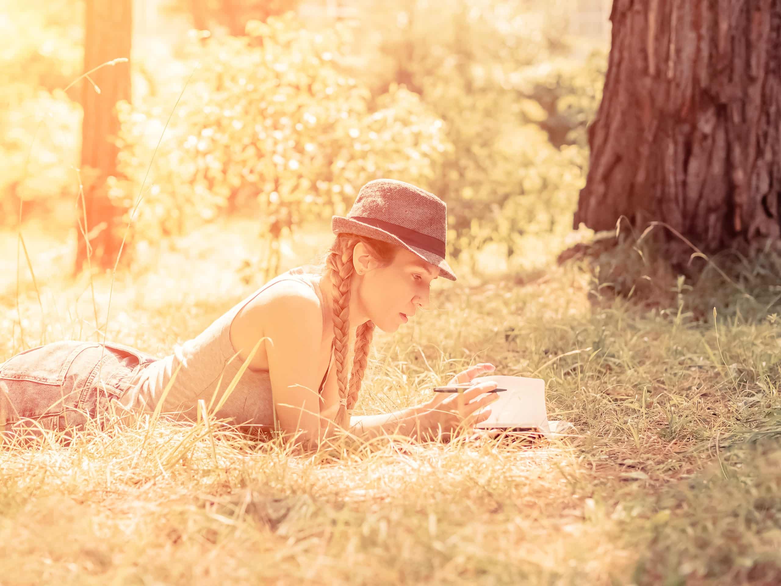 Blonde girl in a hat with two pigtails lies in the grass in a sunny forest writes in her notebook.