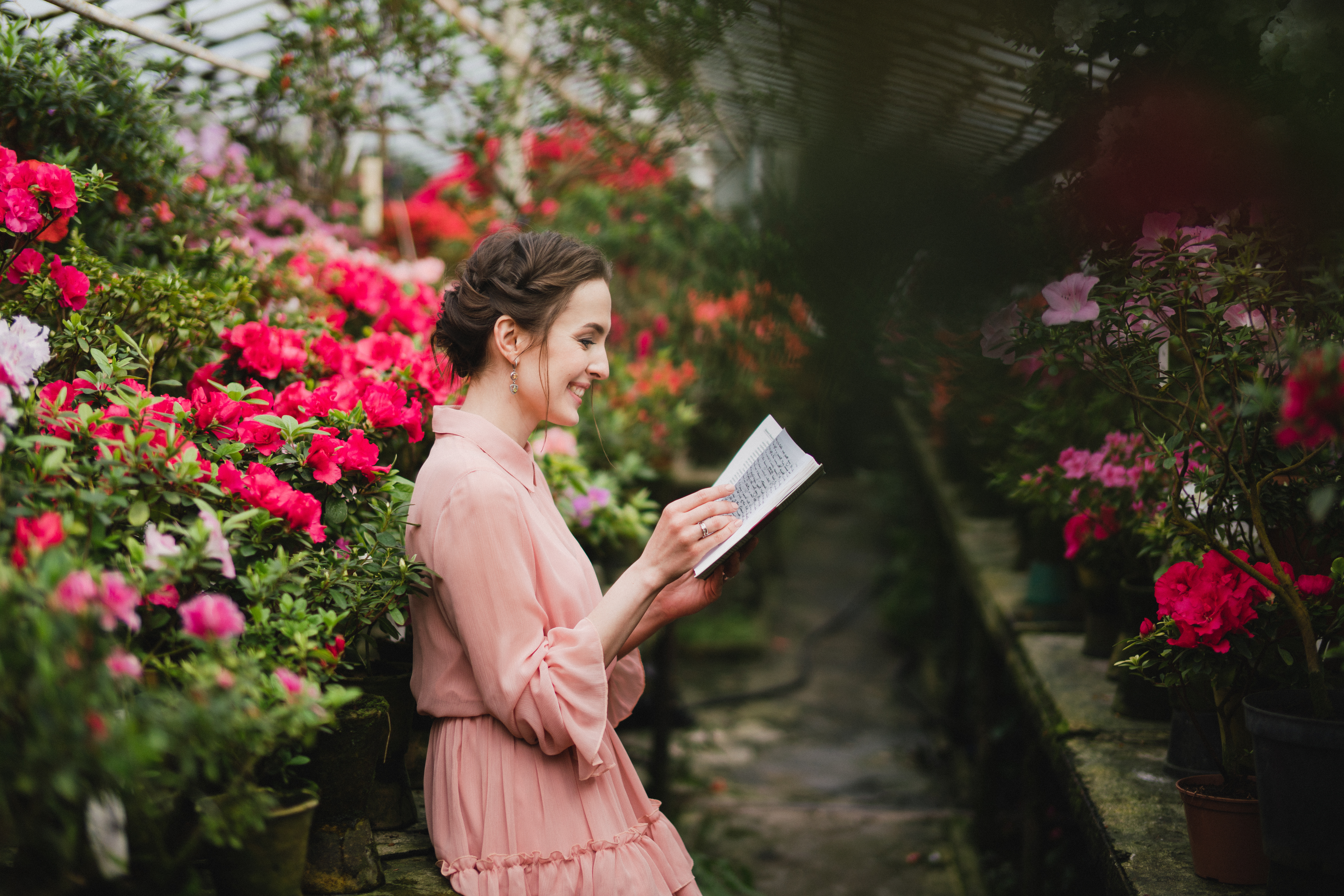 Young beautiful caucasian woman in glass greenhouse among colorful azalea flowers. Art portrait of a girl wearing a romantic pink dress and reading a book.