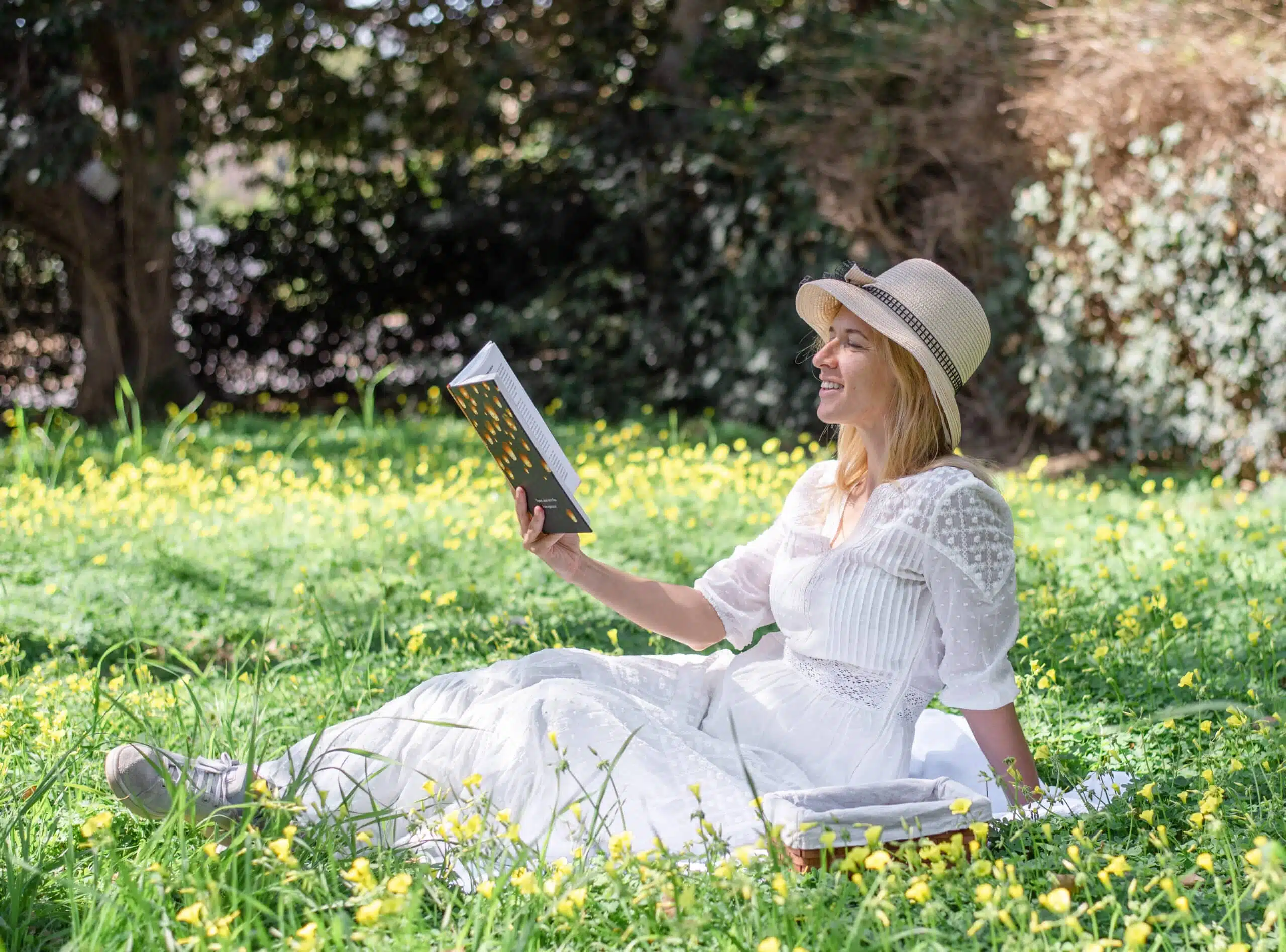 Blonde in white dress and hat sit on green grass holding a book.