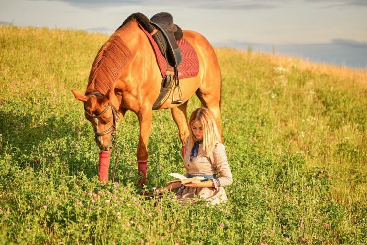Young woman with a book in the field next to her horse.
