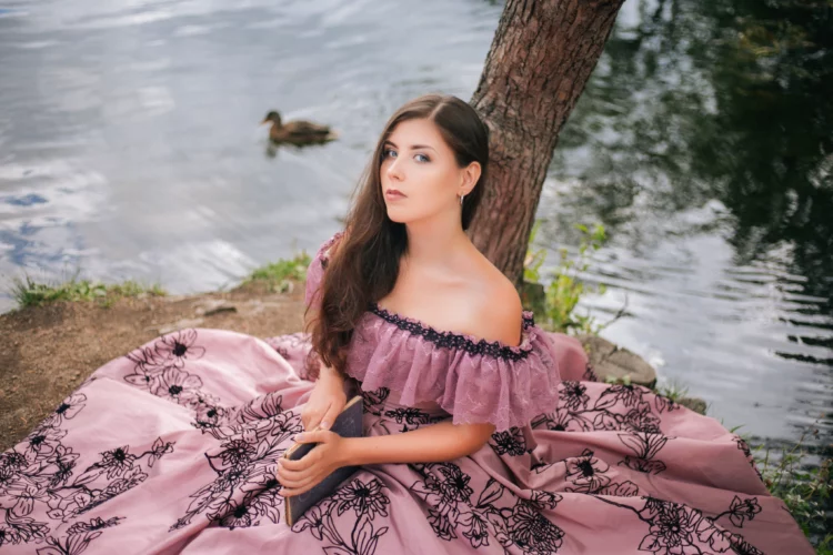 A beautiful young woman in a 19th-century dress sits with a book by the lake.