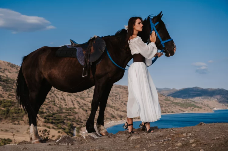 A young lady in a white dress gently hugs her horse against the backdrop of a mountain.