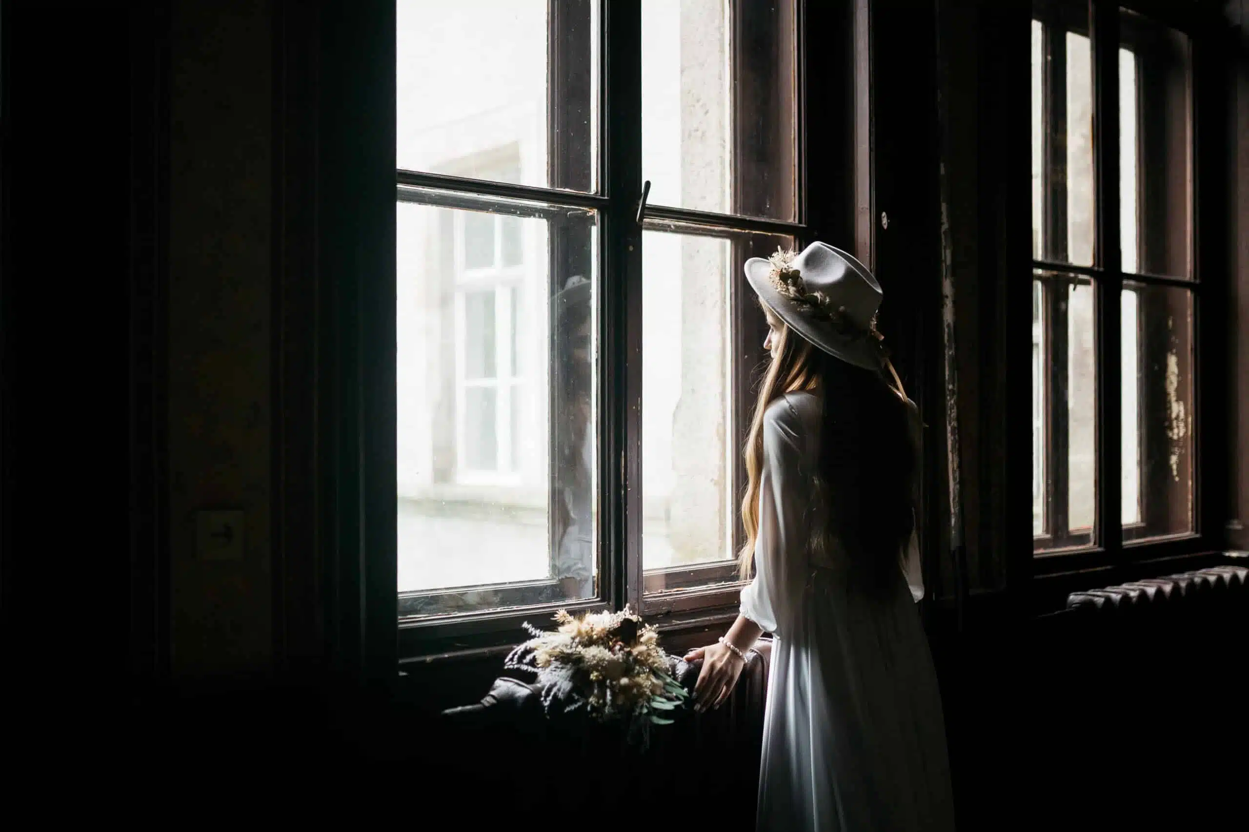 The bride in a hat and a bouquet looking out the glass window.
