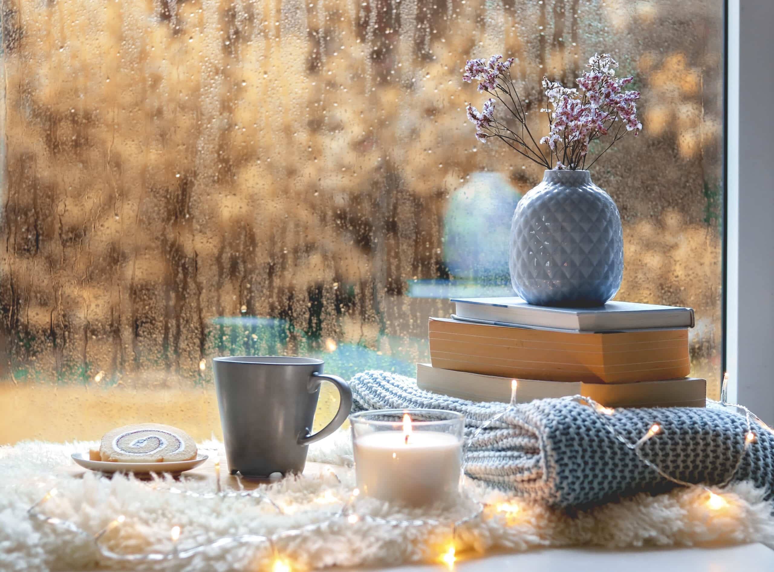 lit candle, a lit candle, flowers in vase on top of stacked books on the window sill, water drops on the glass.