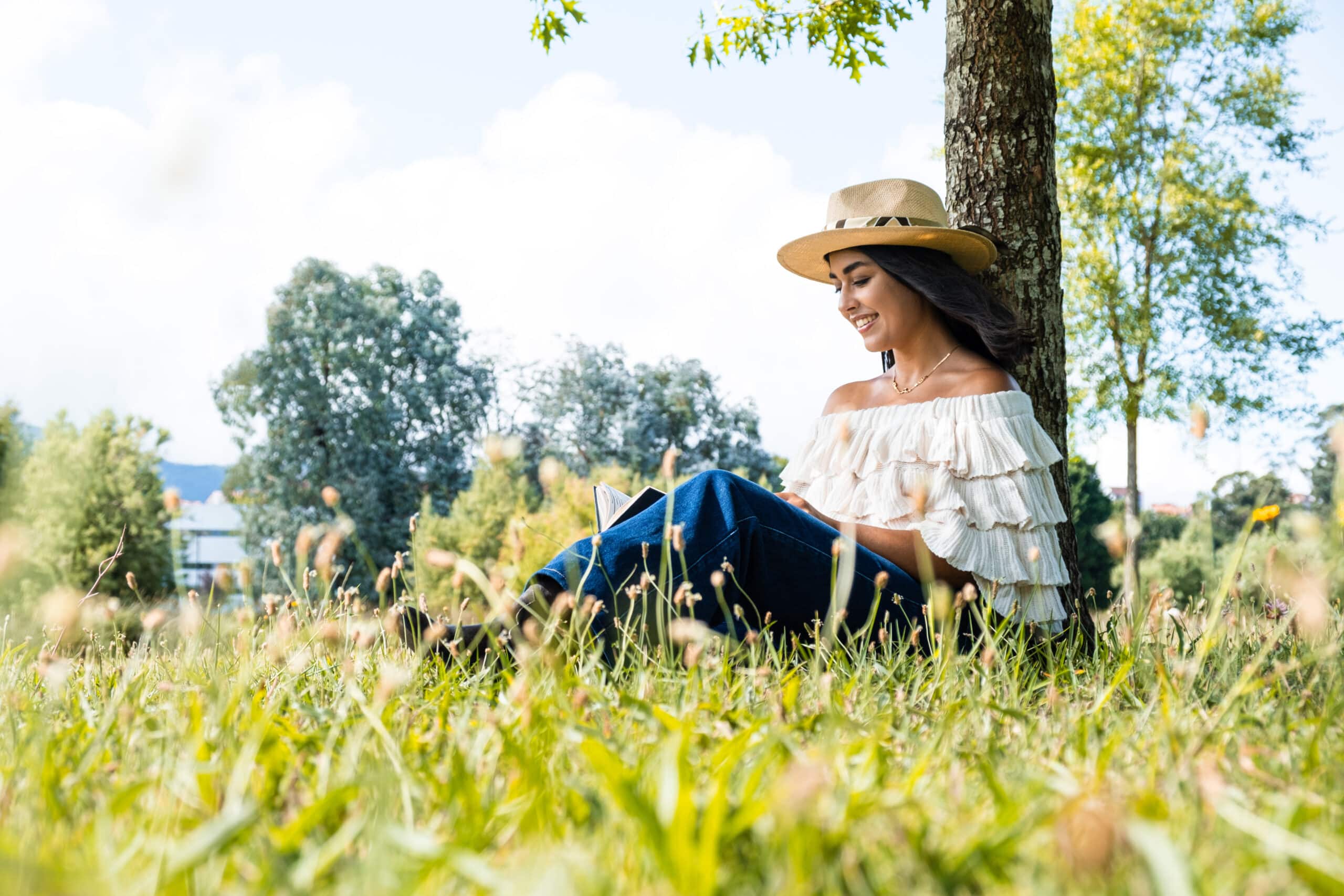 girl wearing a hat and jeans reading a book under the shade of a tree