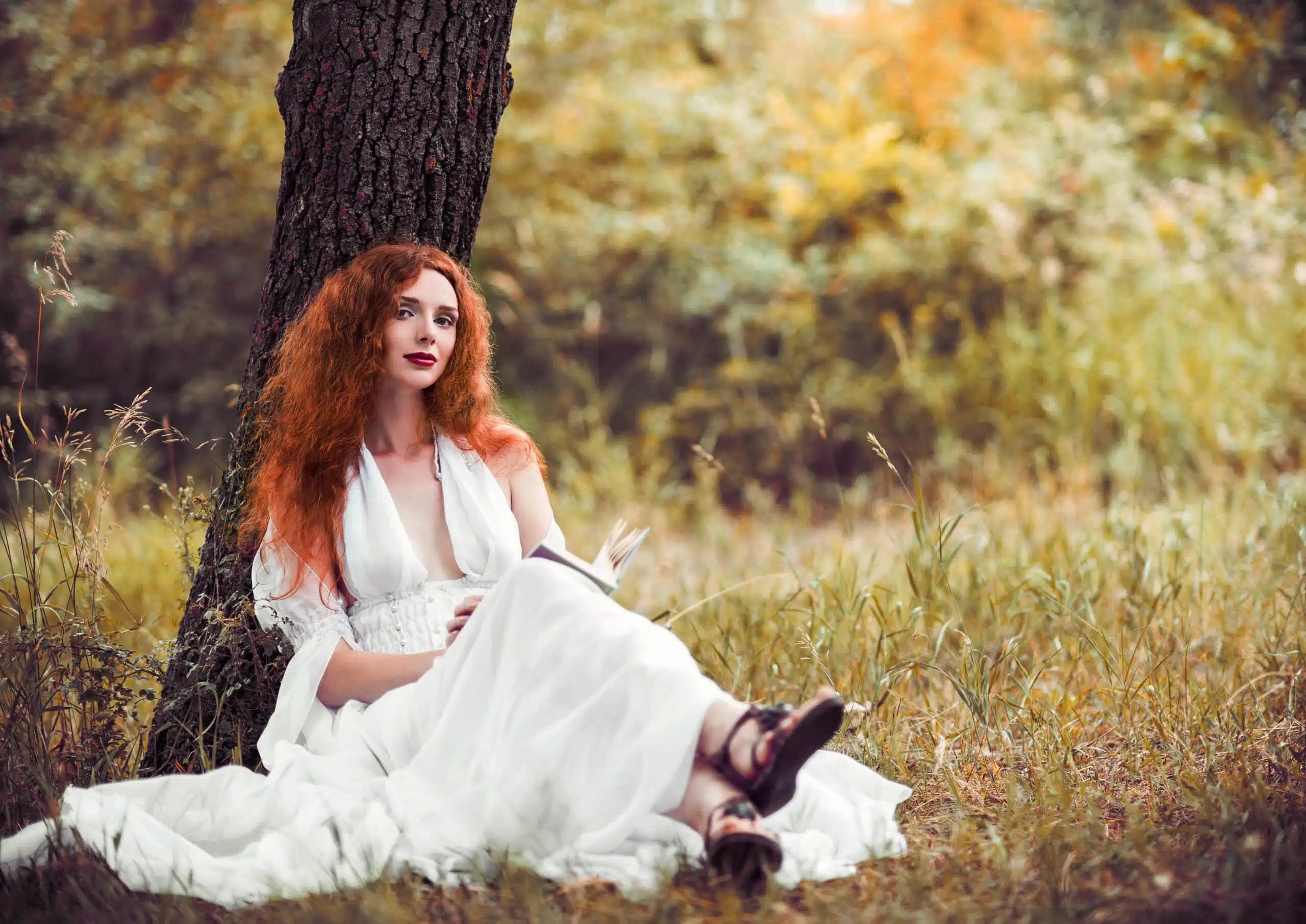 Lovely redhead in white dress woman sitting under tree and reading a book