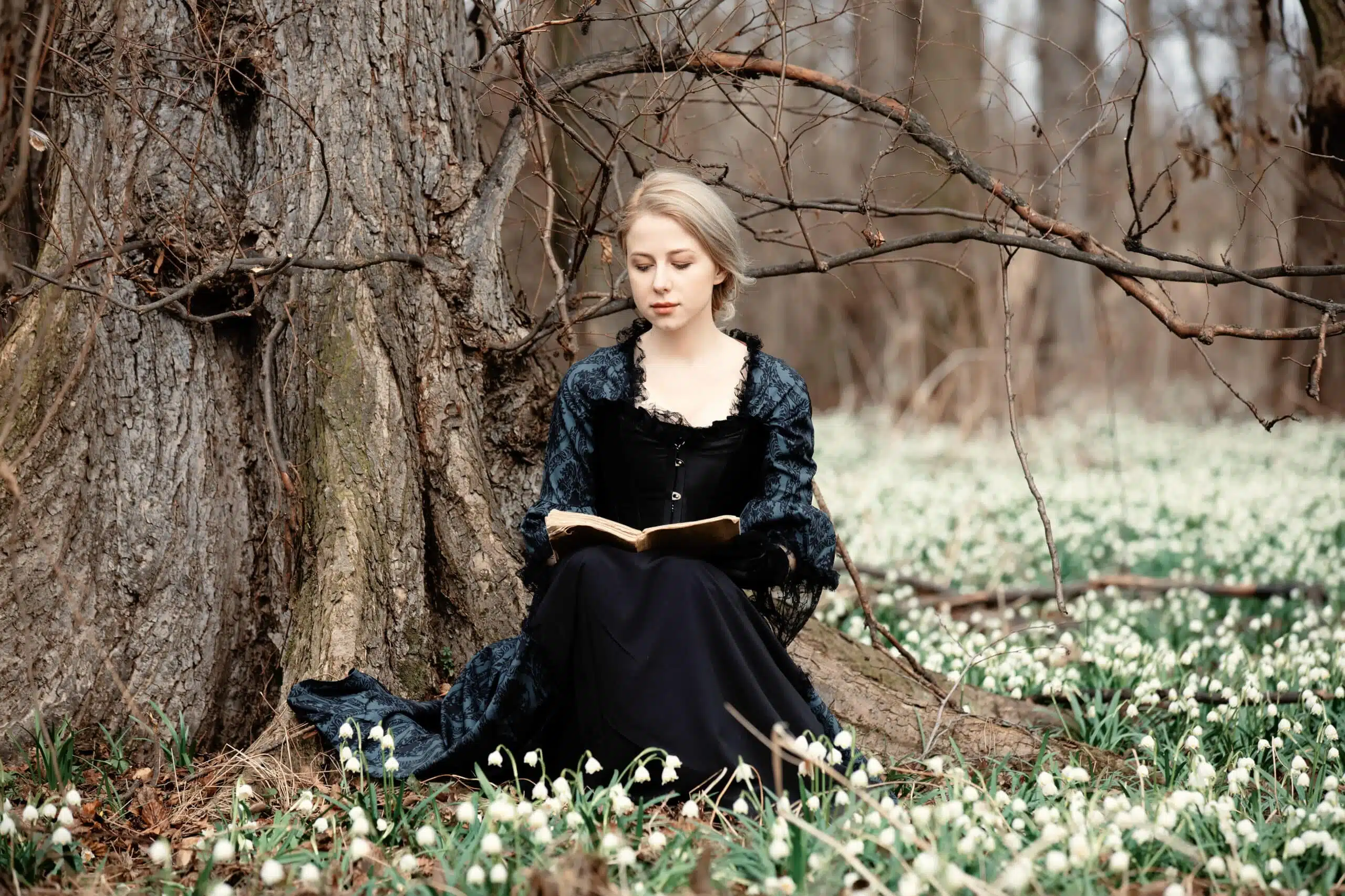 young lady in vintage dress with book is sitting on meadow with snowdrops