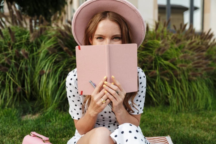 Redhead woman sit on green grass covering face with a book