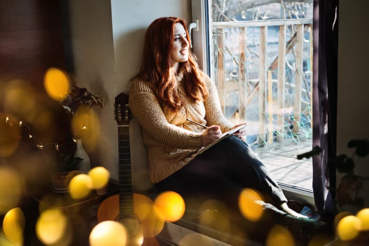 redhead woman sitting on the window sill writing in her notebook