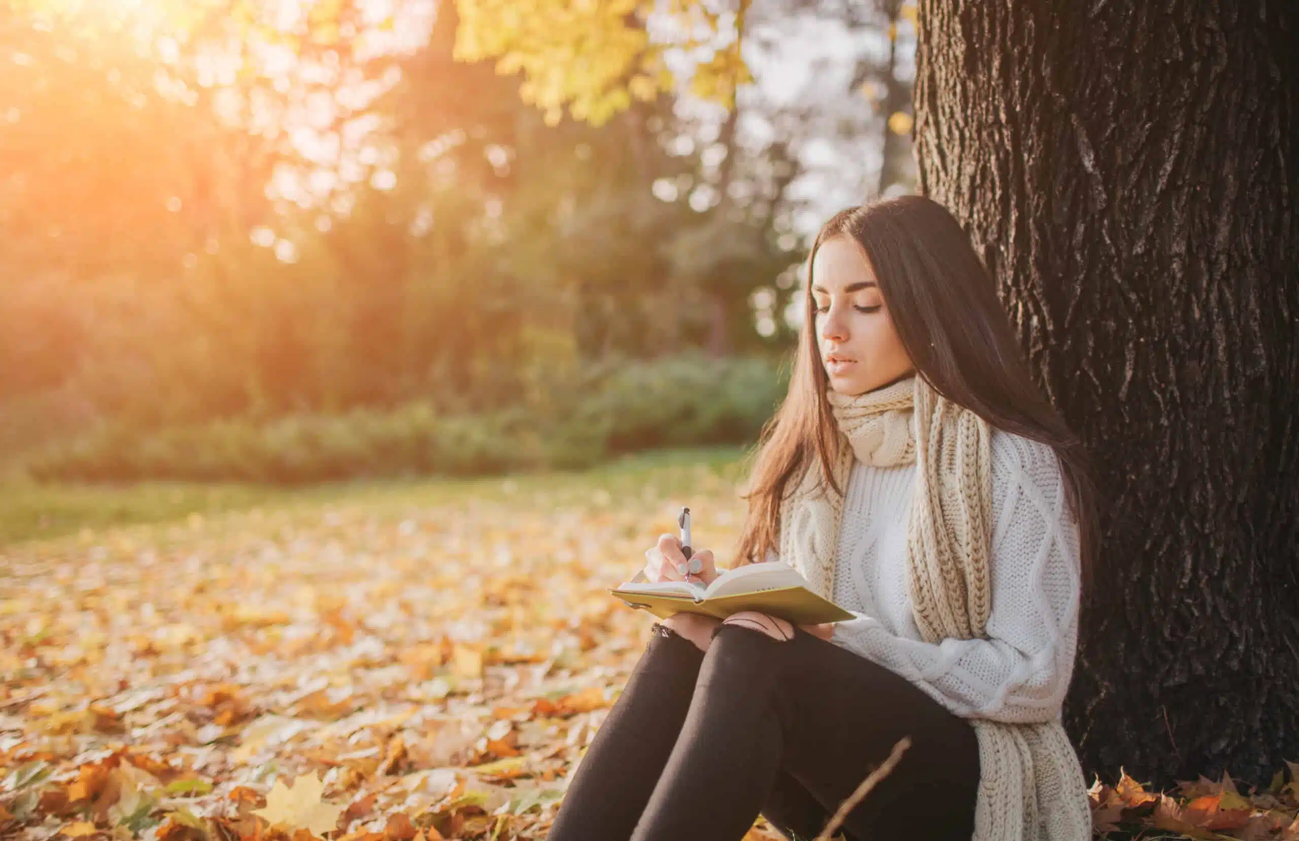 Beautiful young brunette sitting on a fallen autumn leaves in a park, writing in diary