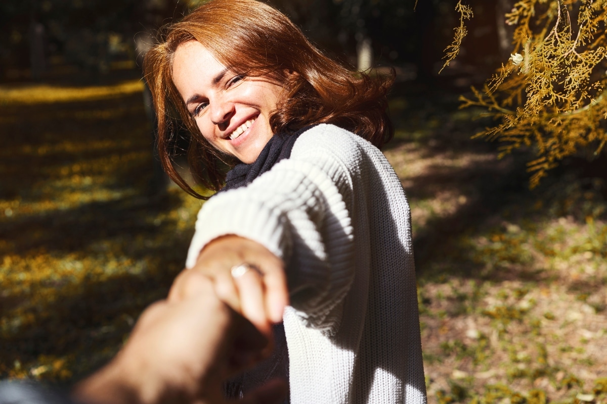 A joyful woman smiling and having fun in autumn park, pulls the hand of her boyfriend and looks at camera.