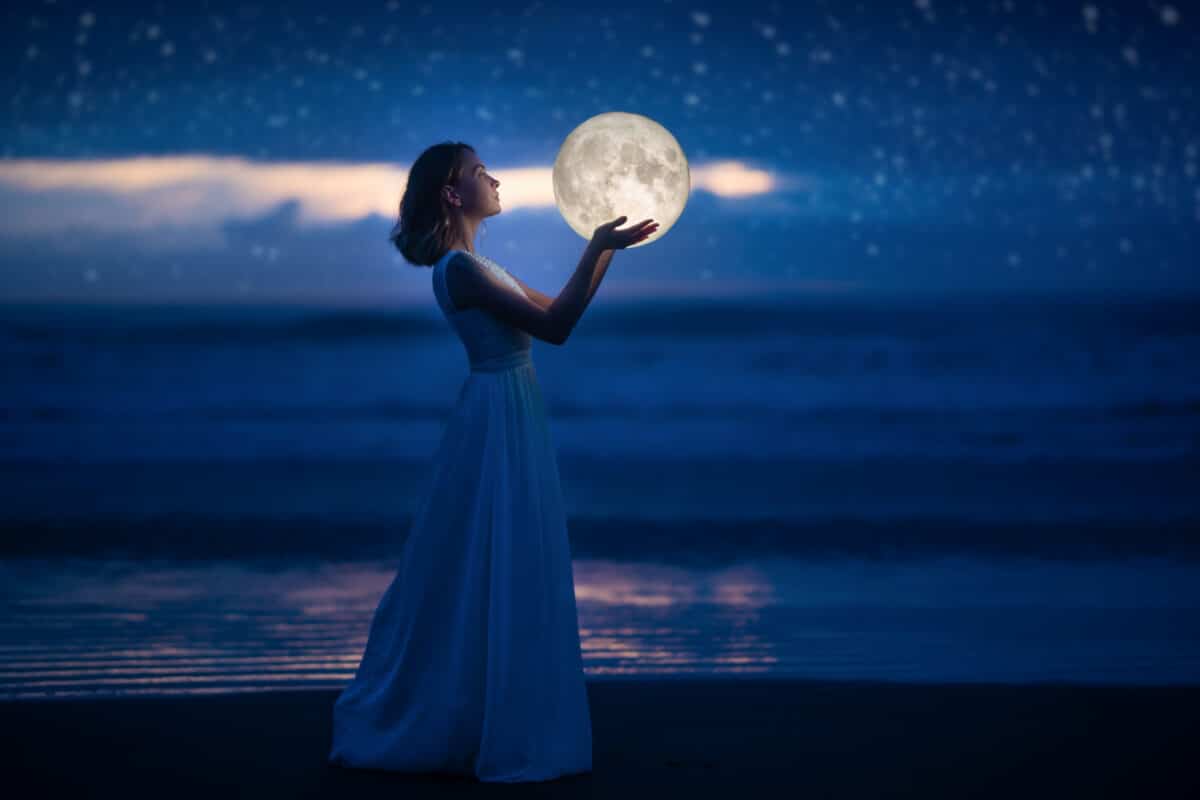 A young girl on a night beach holds the moon, with a starry sky.