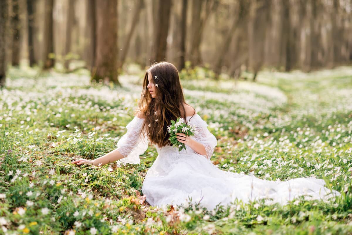 young beautiful woman in a white dress collects primroses in the spring forest and holds a bouquet of white anemones in her hands