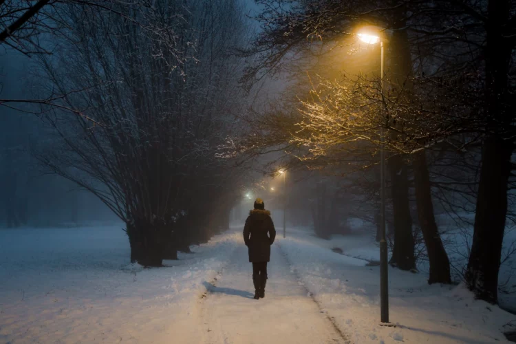 Young woman alone slowly walking on sidewalk under yellow lights in winter.