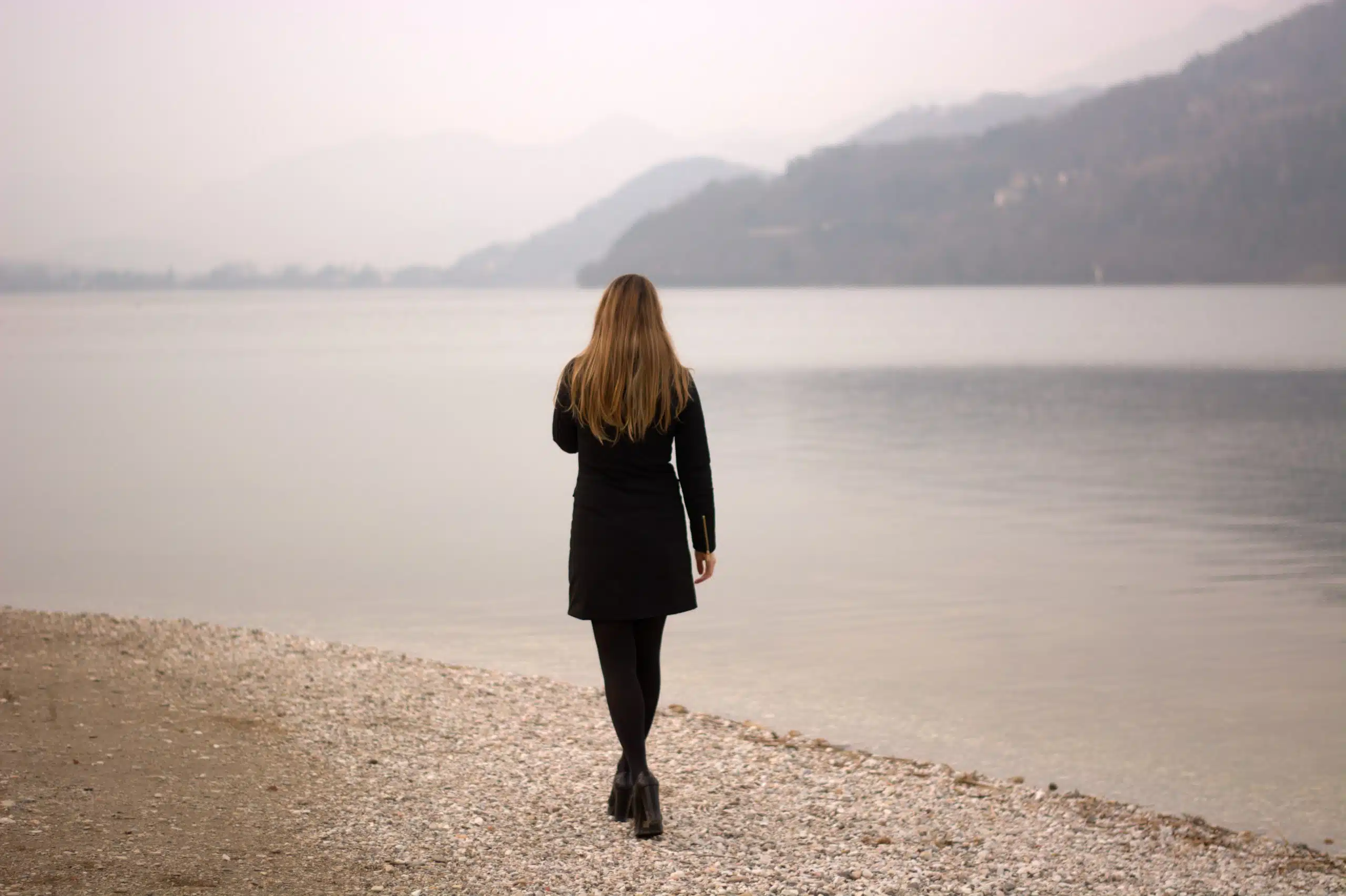 Melancholic woman in black walking alone by the foggy lake.