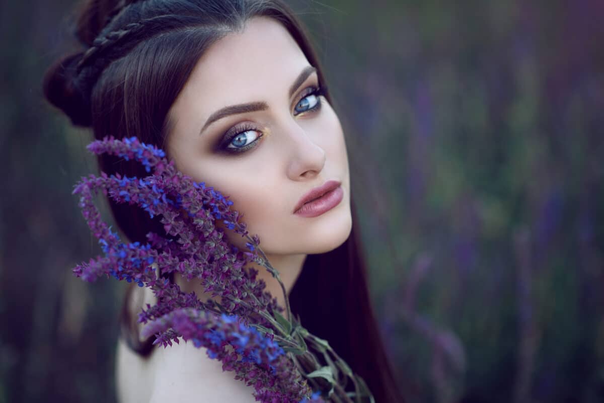Close up portrait of beautiful blue-eyed lady with perfect make up and plaited hairstyle sitting in the field and holding purple and blue flowers at her face. Blurred background.