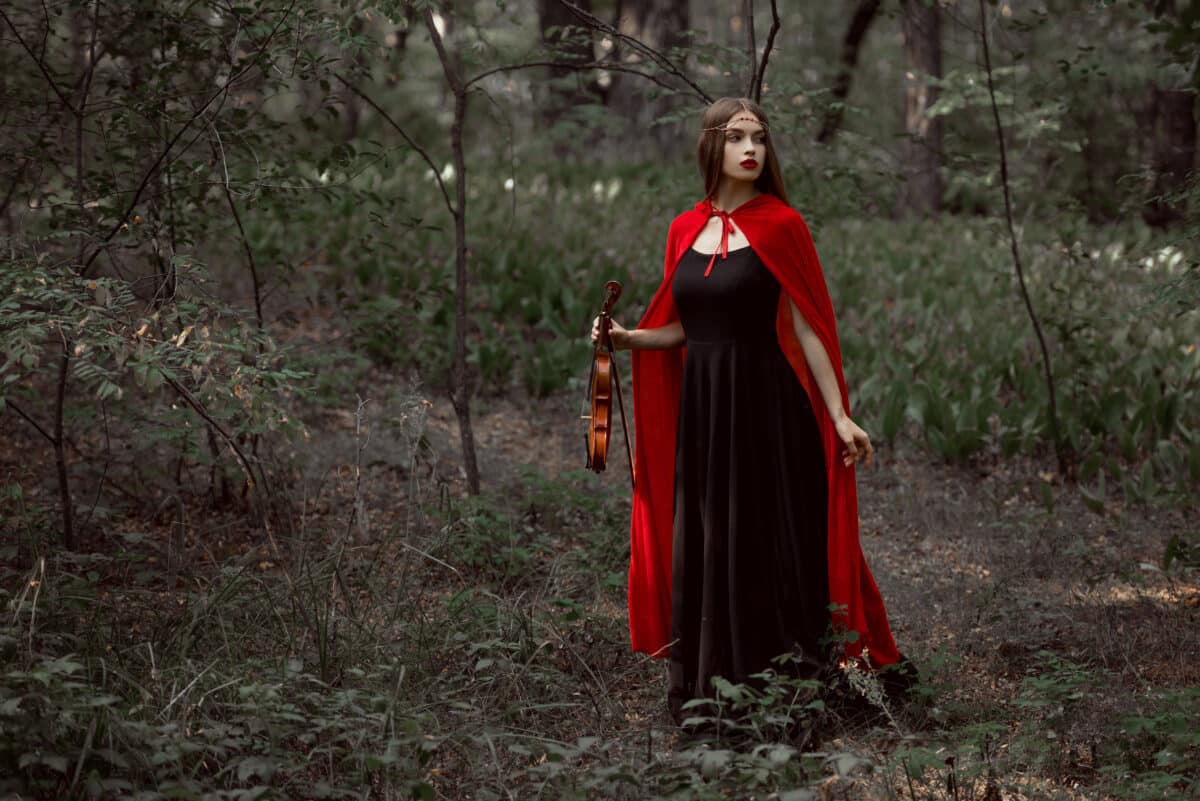 beautiful mystic girl in black dress and red cloak holding violin in forest