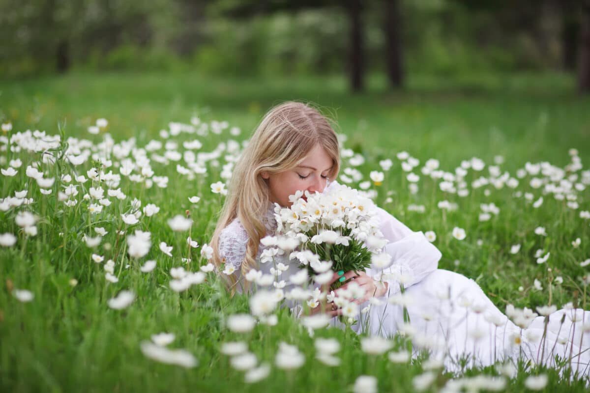 a pretty blonde in a vintage white dress rests on the grass with white anemone flowers