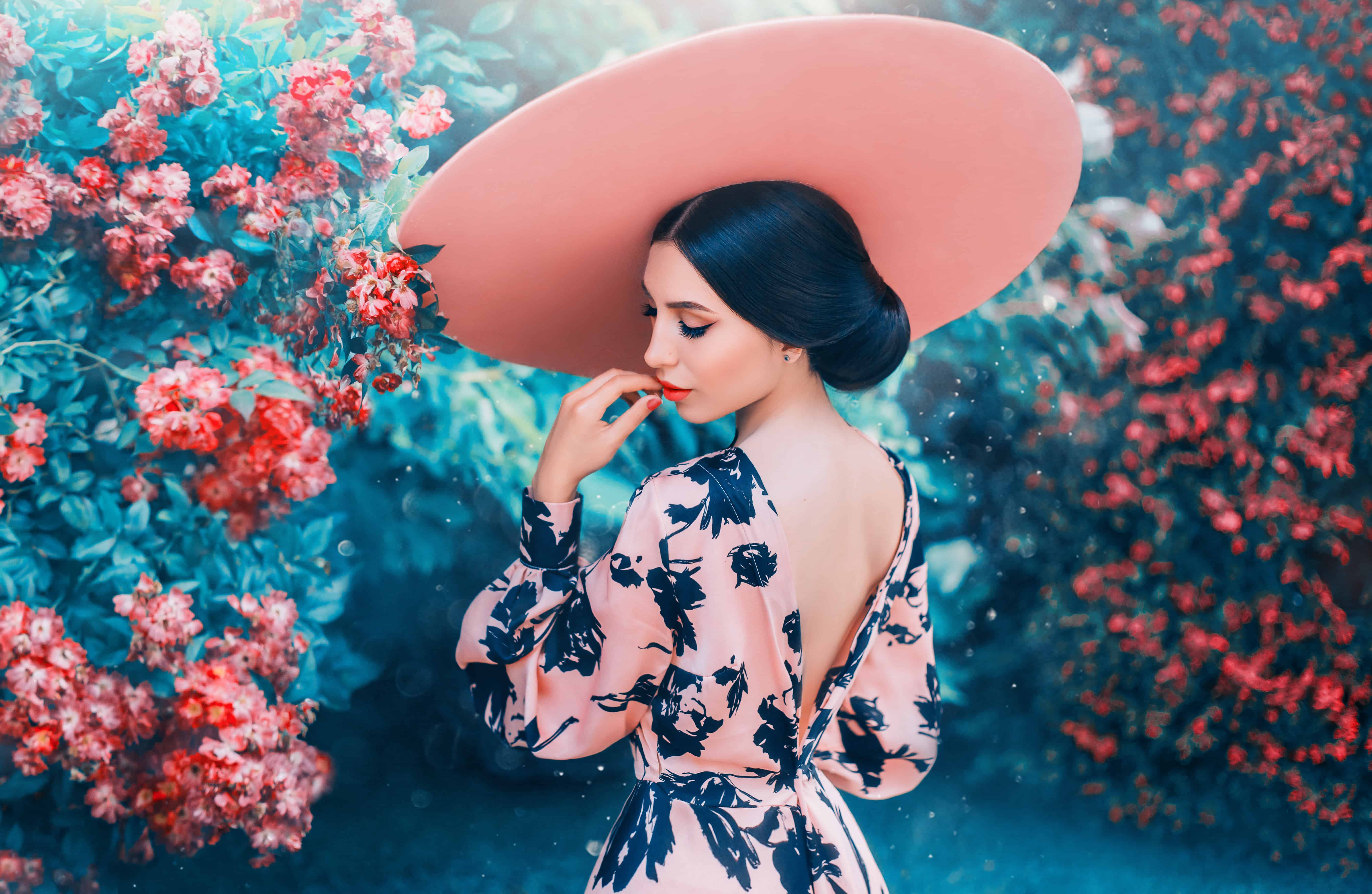 cute woman with perfect gentle hairdo from long black hair wearing pink hat with wide brim, elegant look for date and photo shoot, lady with open back and bright lips, girl in blooming roses