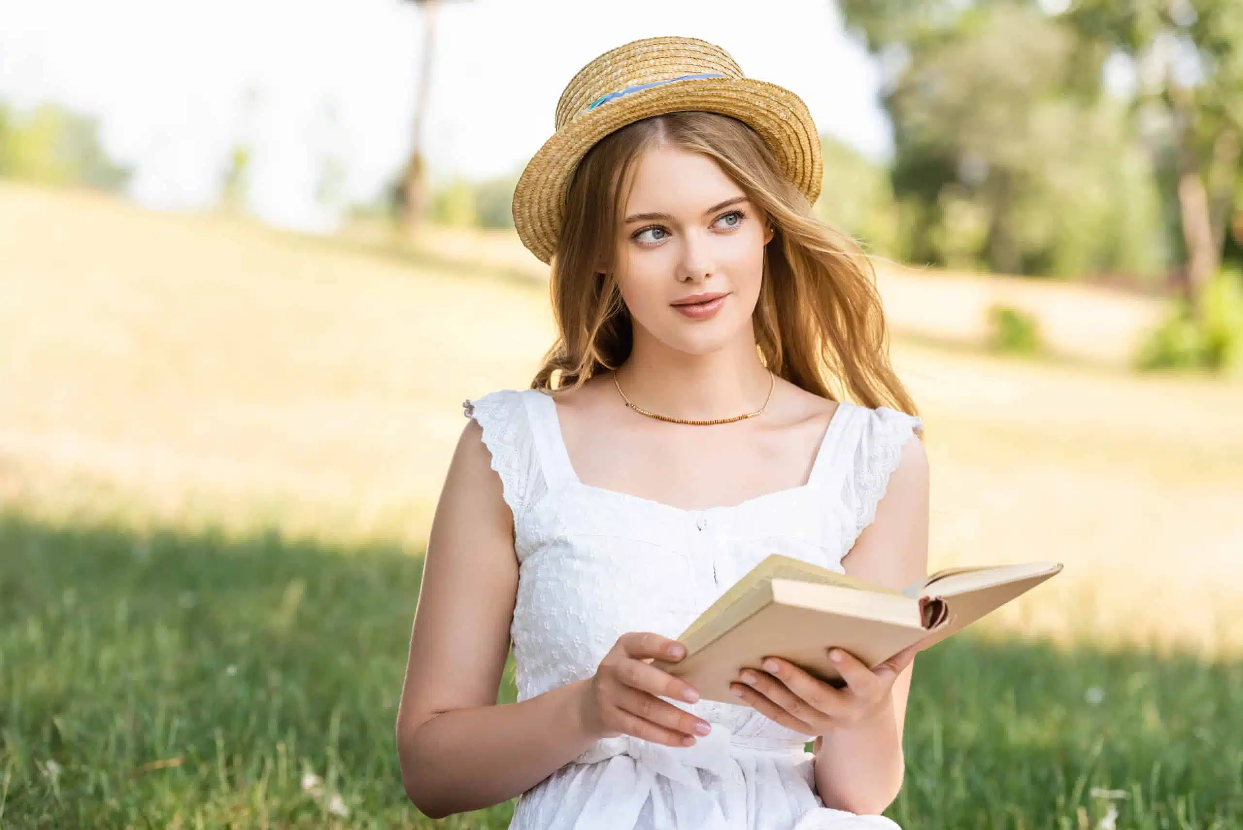 Beautiful woman in white dress and straw hat holding book while sitting on a meadow.