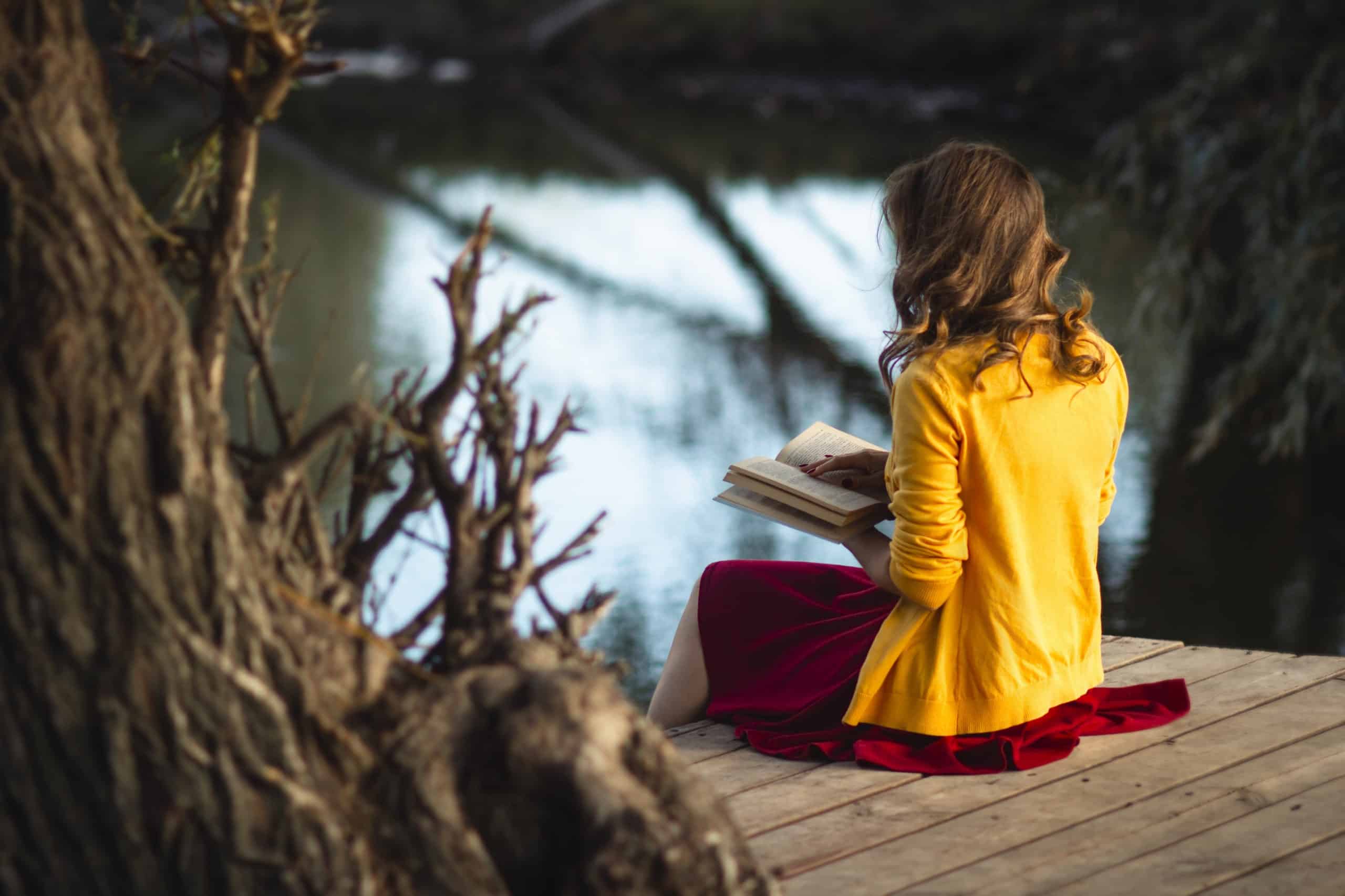 Beautiful young woman on the river banks sitting on a wooden platform reading a book.
