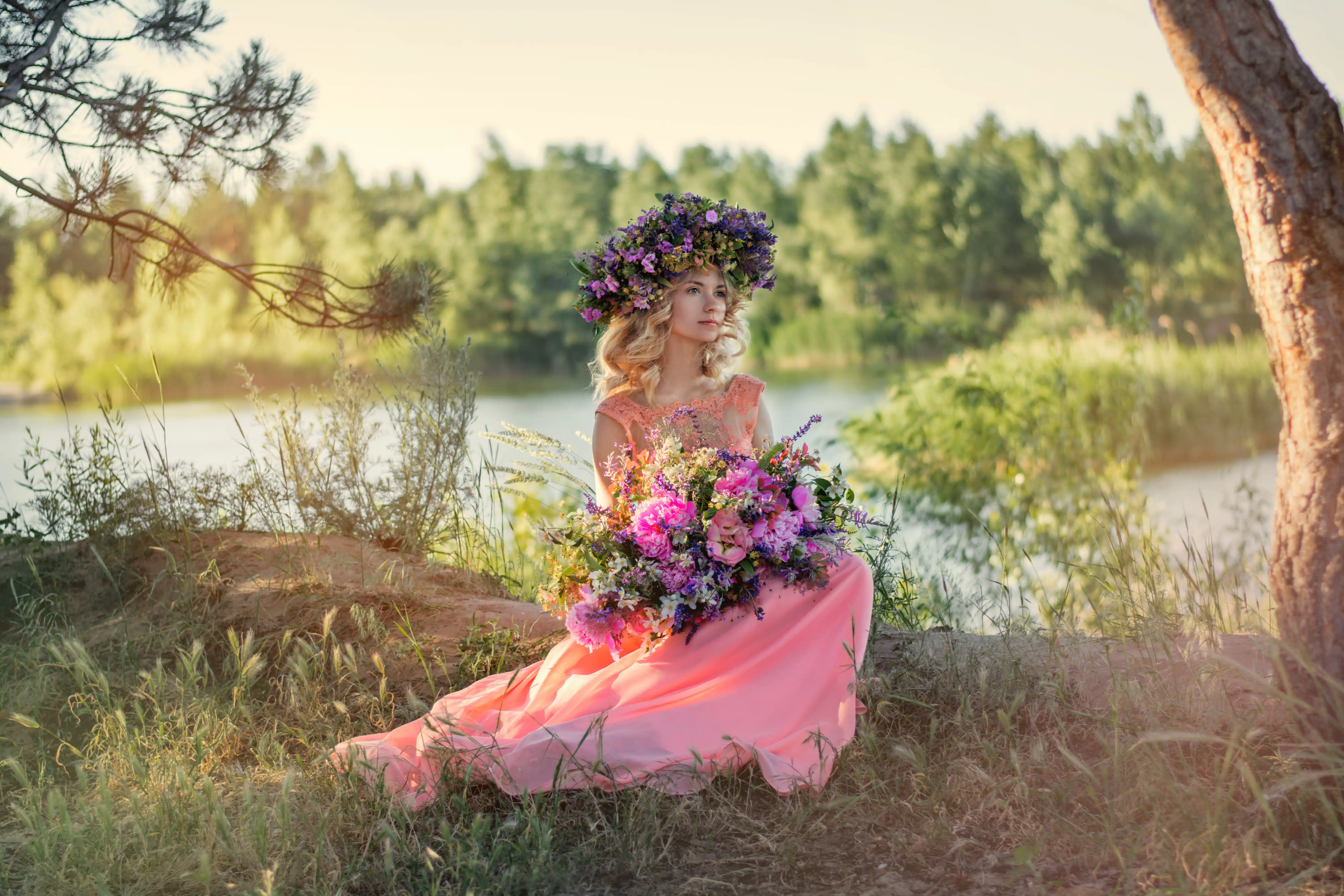 lady in pink dress and flower head wreath holds a big bouquet of flowers sits by the river