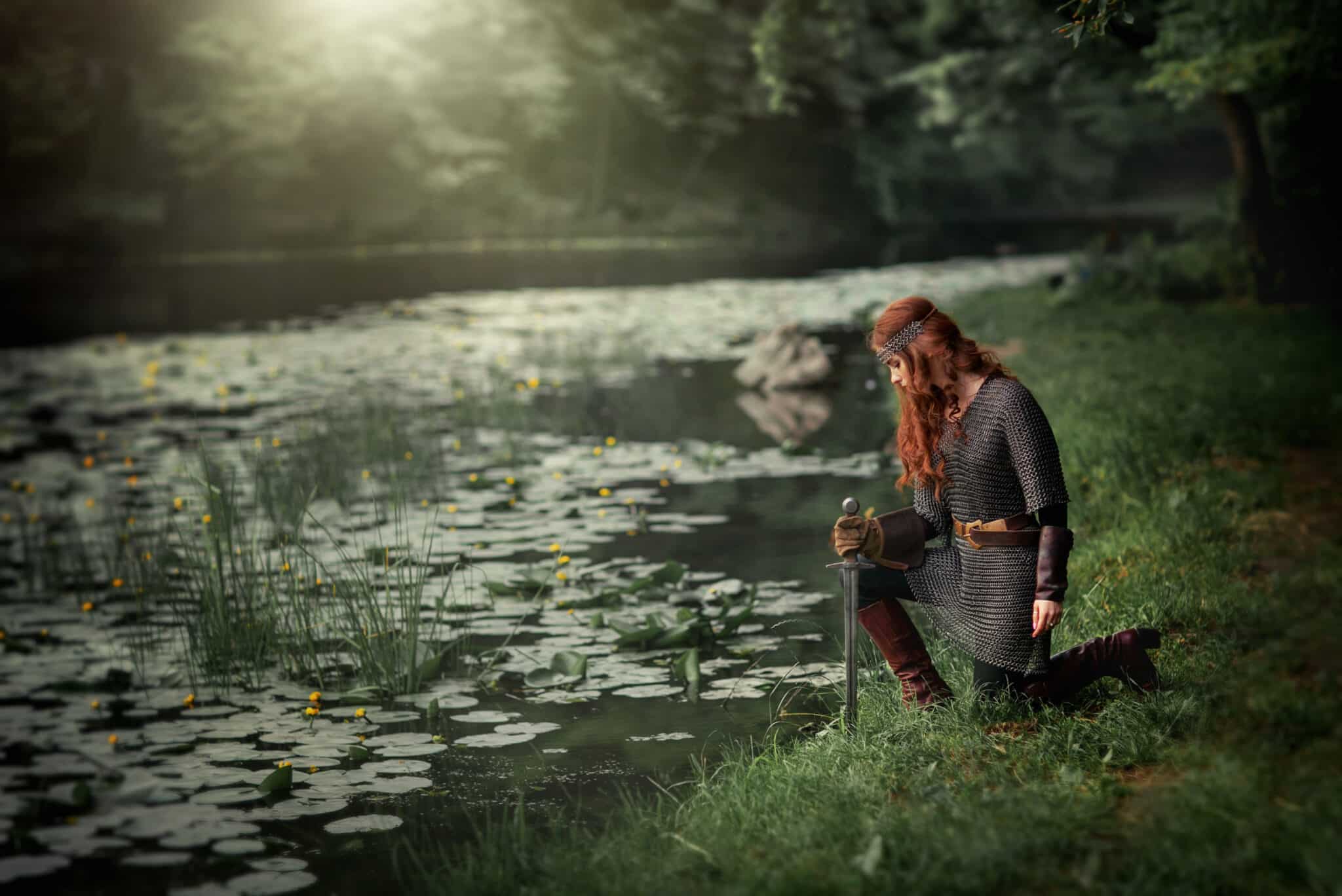 Beautiful red haired woman in metal medieval armor kneeling down by the lake with a sword