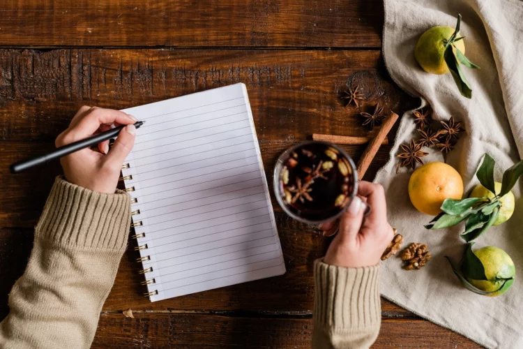 Girl hands holding glass of mulled wine and pen over blank page of notebook.