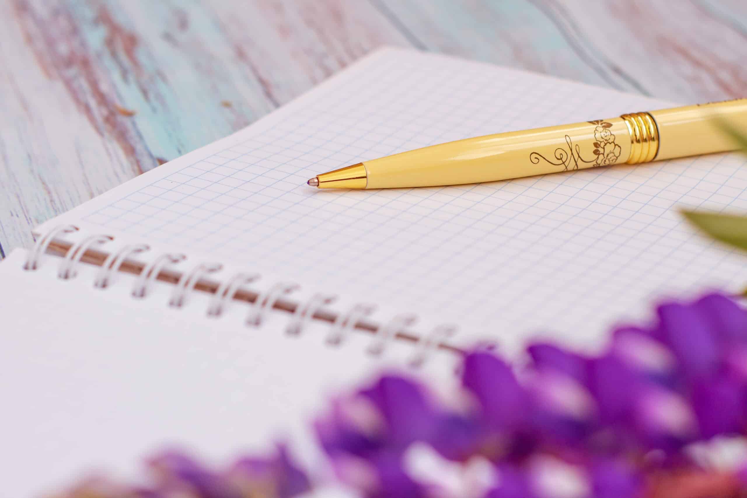 Notebook with a yellow pen, next to purple flowers.