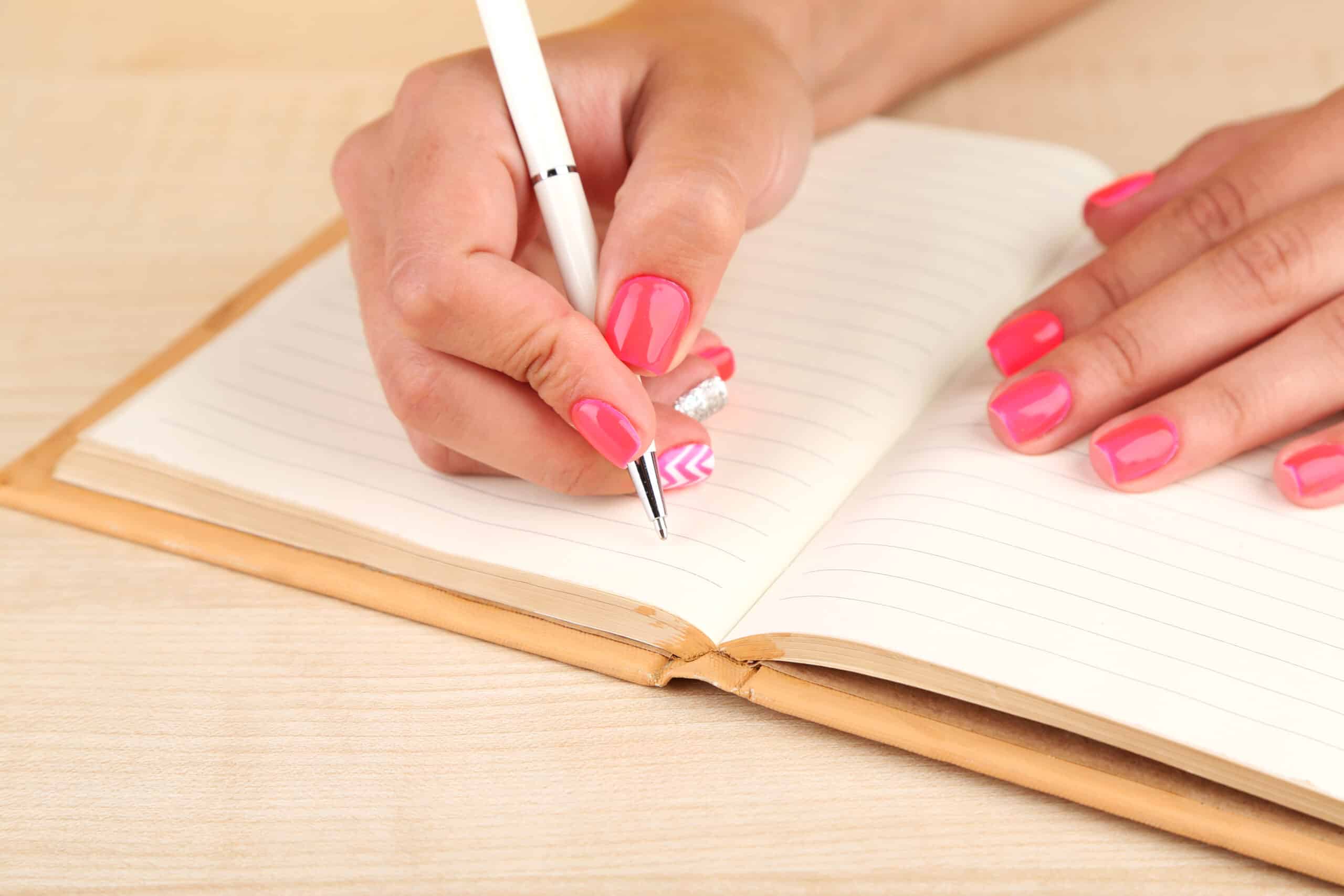 Female hand with stylish colorful nails witing in notebook with pen.