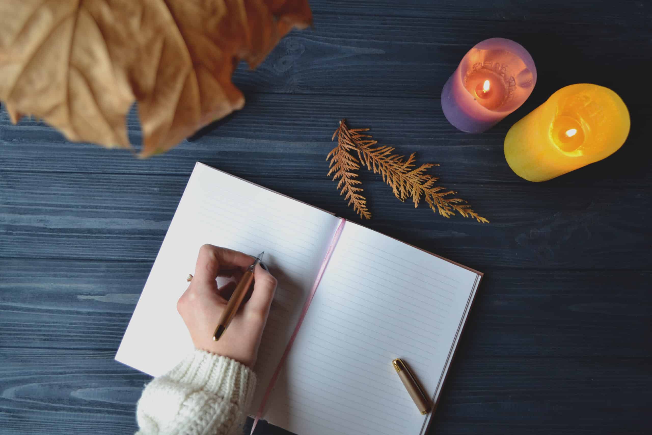 Woman's hand writing in the notebook on aesthetic wooden desk with lit candles.