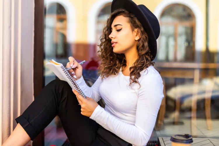 Attractive young woman writing into notebook while sitting on bench in the park