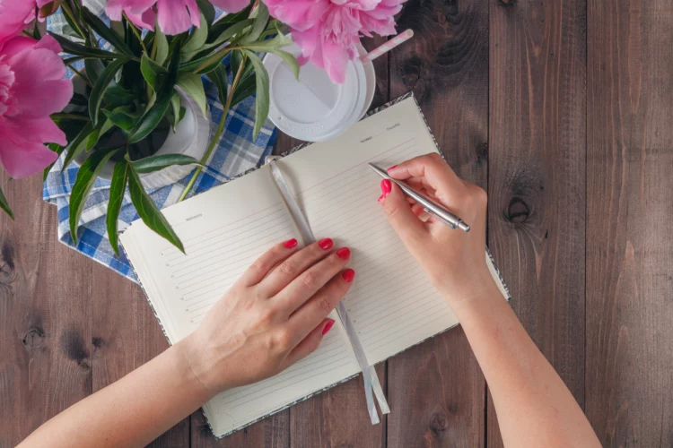 female hand with red nail polish writing on blank notebook, with a vase of pink flowers