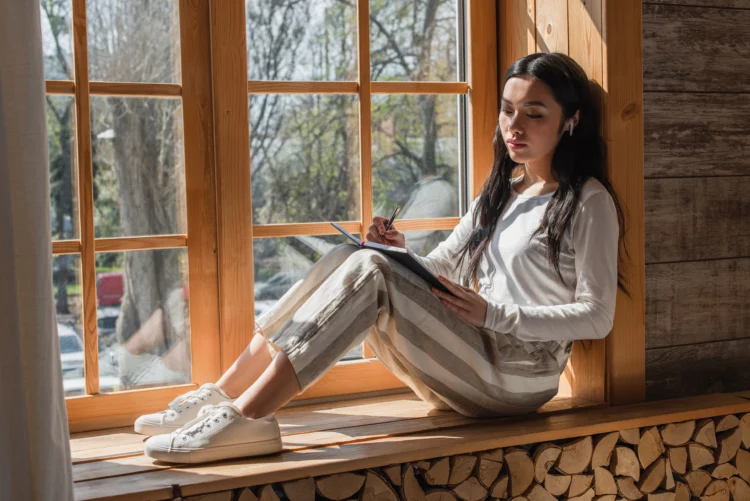 dreamy young woman in casual clothes writing sitting on cozy window sill.