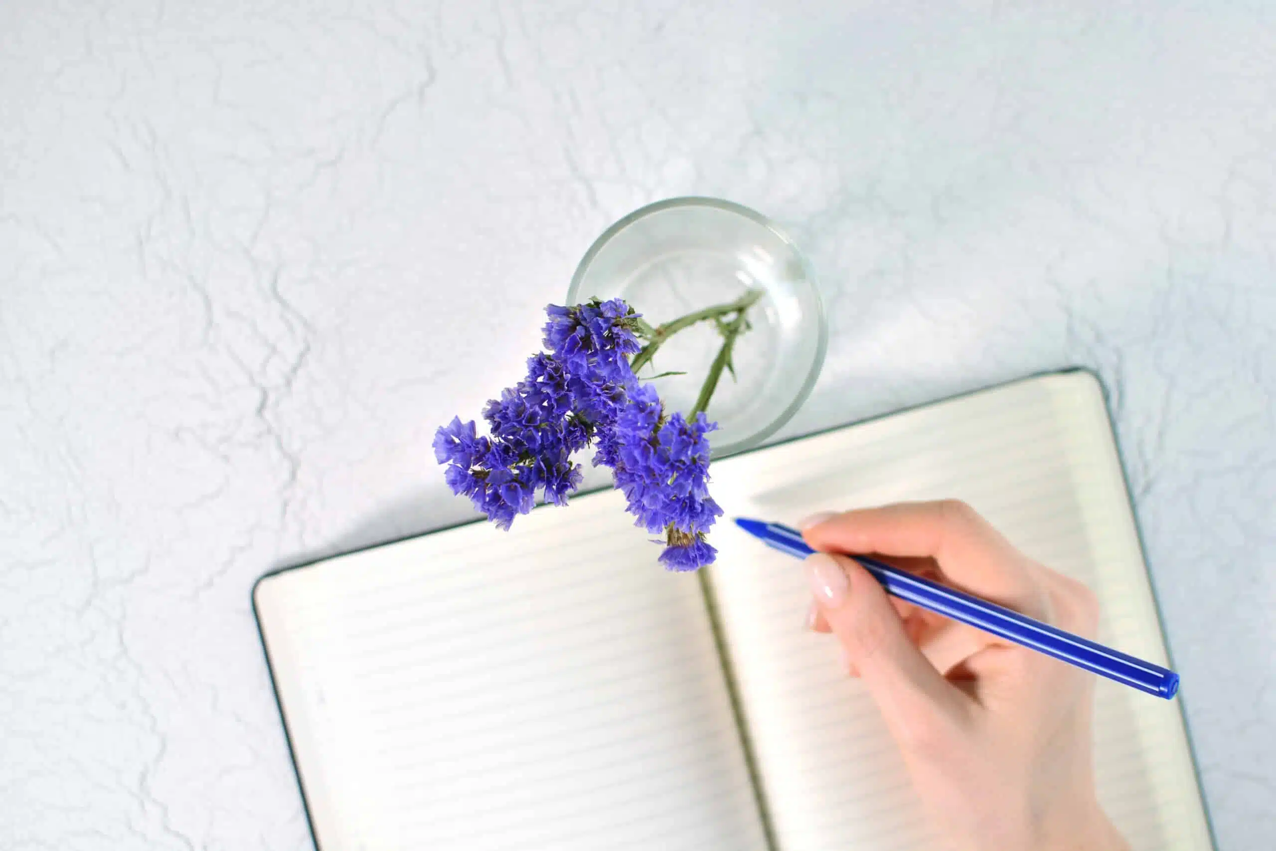female hand with pen above the notepad and a glass with purple flowers.
