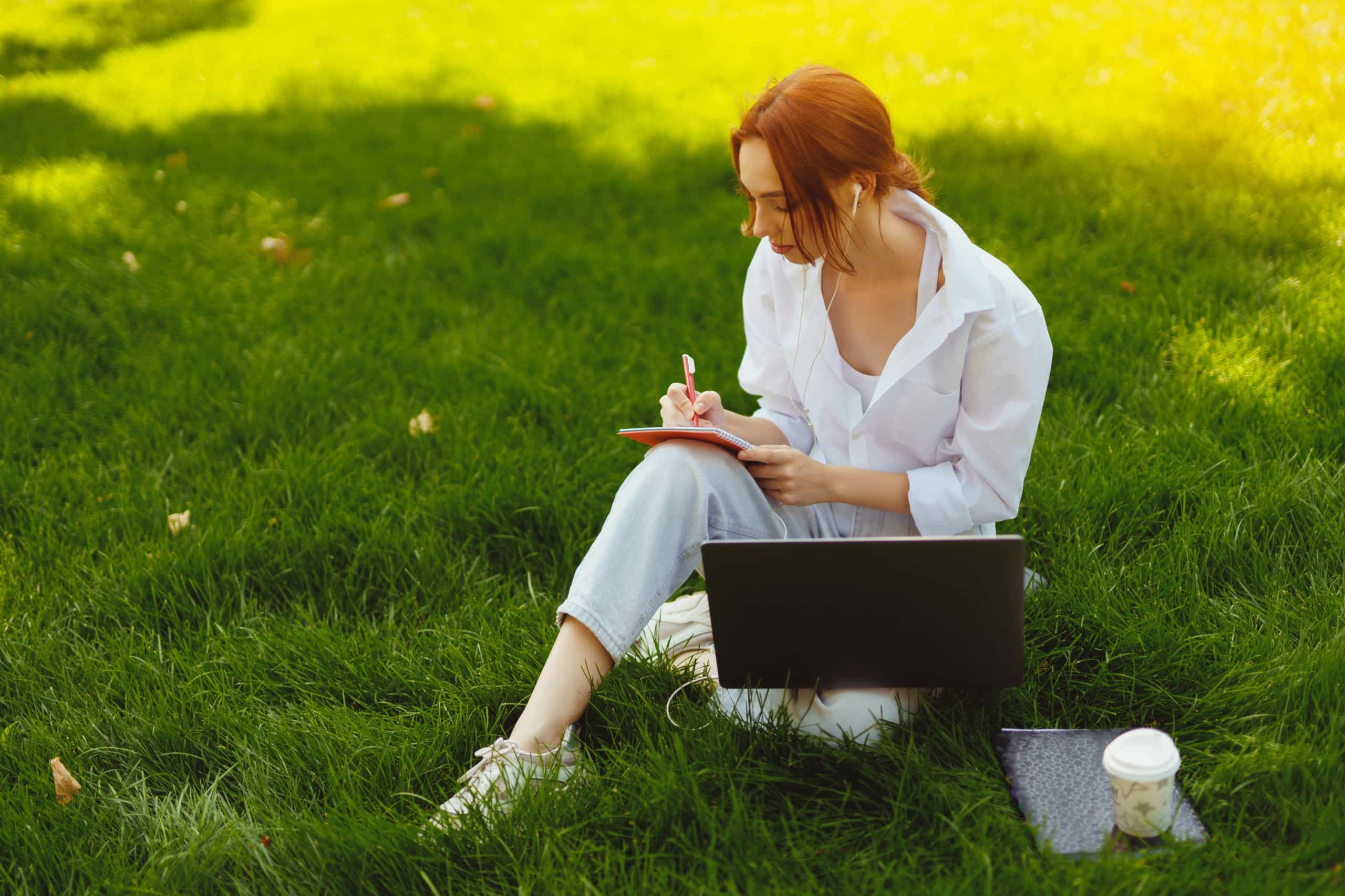 Beautiful young redhead in park outdoors writing in her notebook, while seated on the grass.