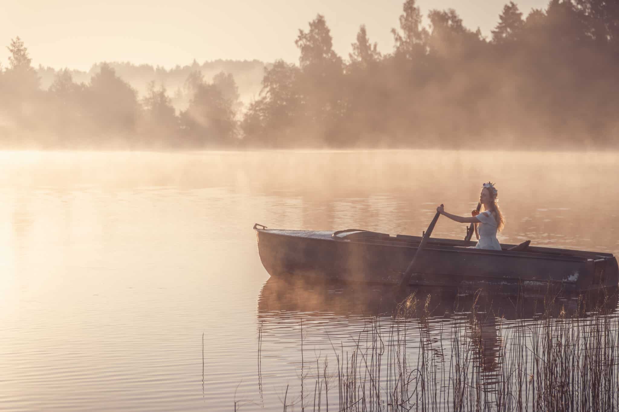 Beautiful lady in a light-blue dress rowing a boat on a misty lake